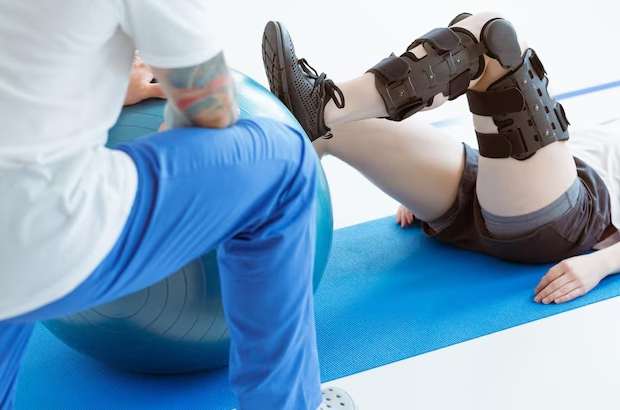 Paciente herido con un soporte para la pierna haciendo ejercicio en una alfombra azul en una oficina de fisioterapia