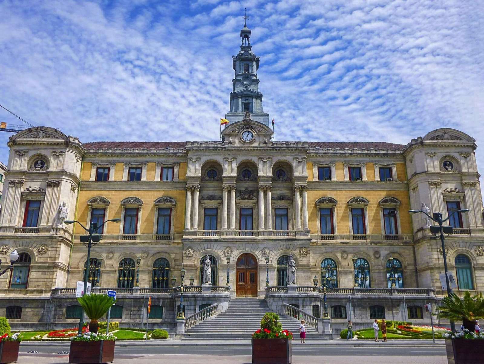Fachada del Ayuntamiento de Bilbao con su torre y reloj, escalinata, jardines con flores y cielo con nubes.