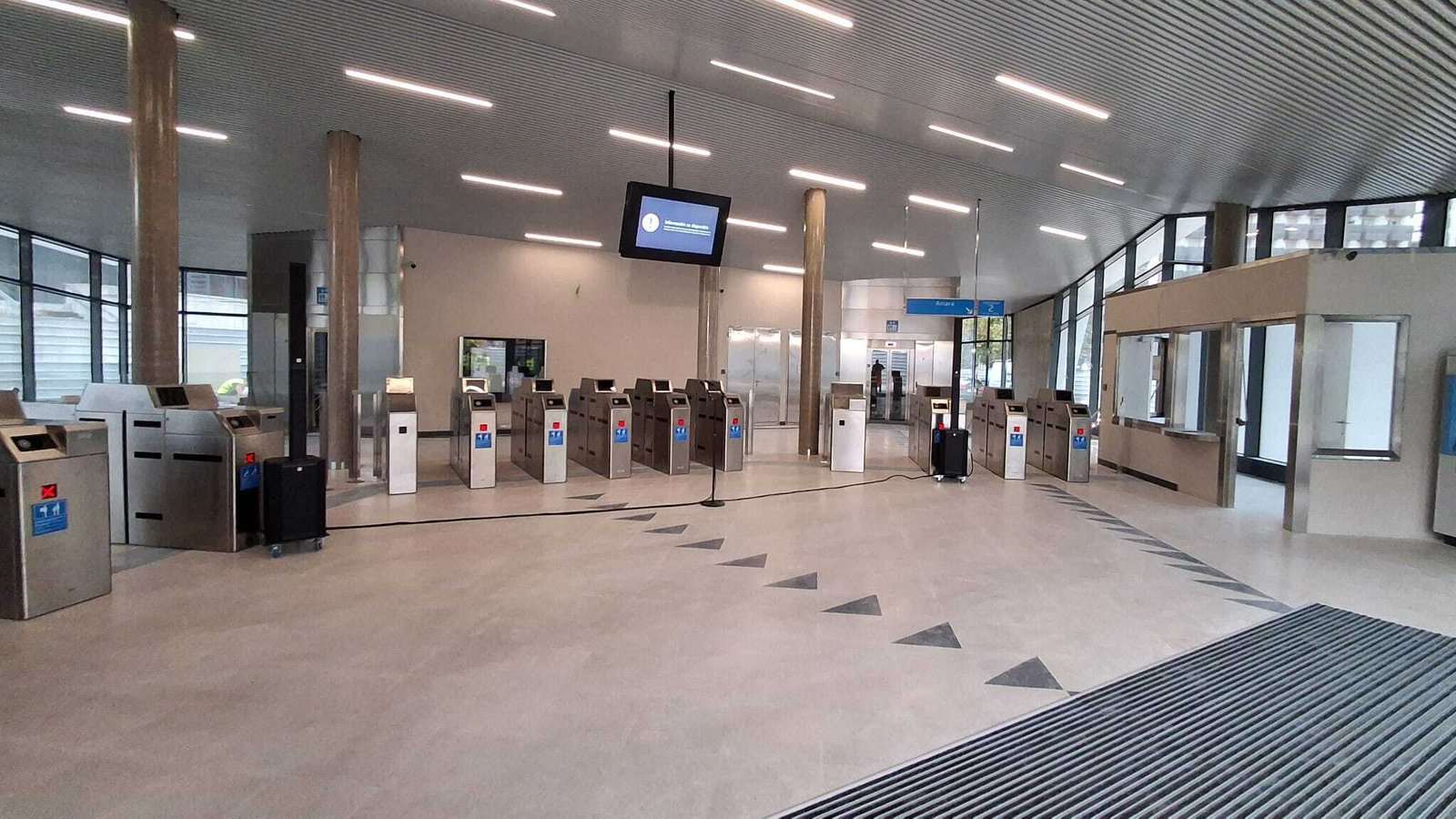 Vestíbulo de estación vacío, con torniquetes de control de acceso, columnas, fachada acristalada y una taquilla