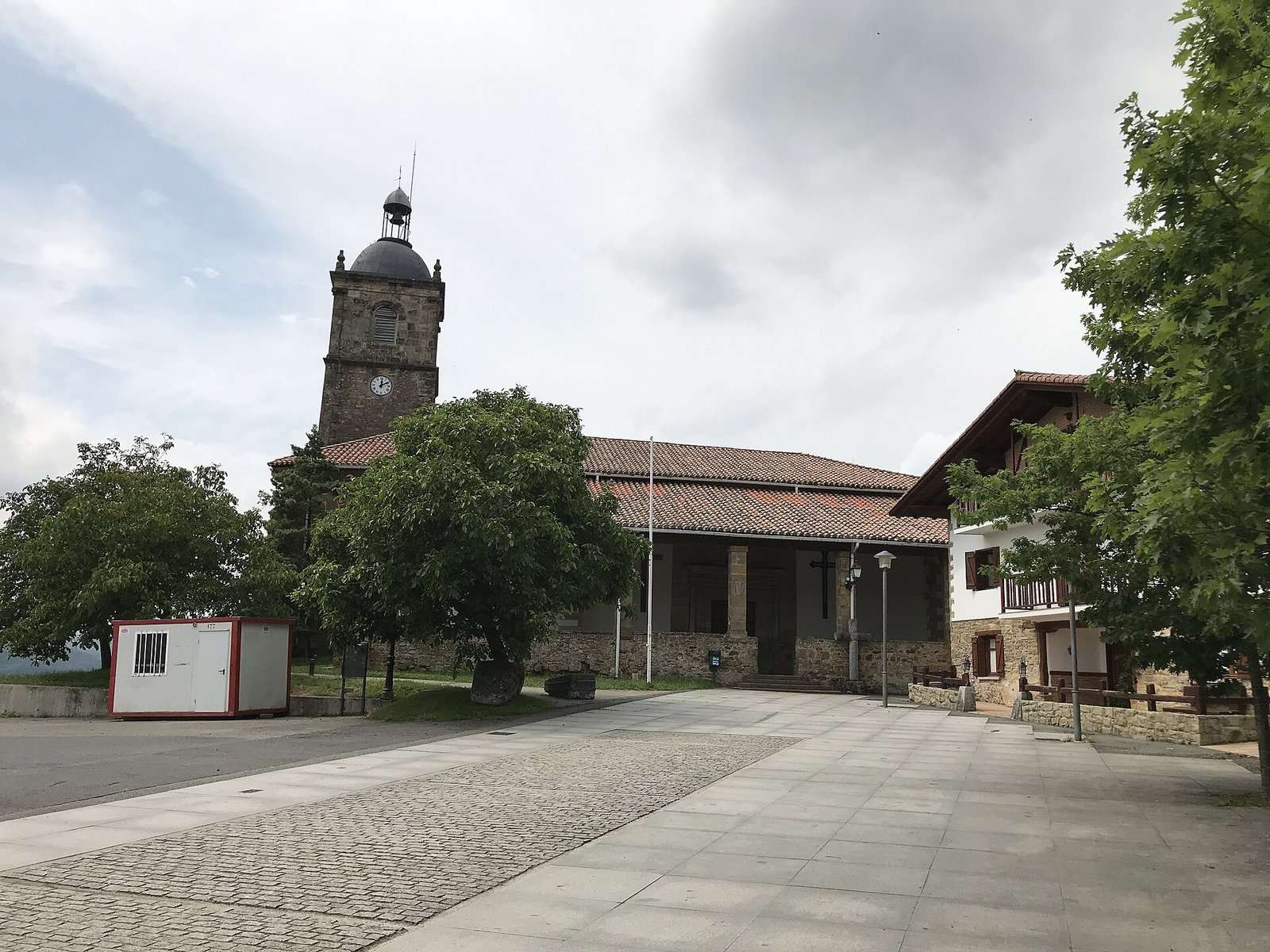 Iglesia de San Pedro de Lumo con torre con reloj y pórtico, junto a una plaza empedrada y un caserío, en Gernika-Lumo.